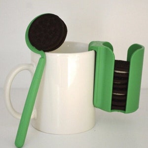 Può includere: Una tazza di ceramica bianca con un porta-biscotti in plastica verde attaccato al lato. Il porta-biscotti contiene una pila di biscotti al cioccolato. Uno strumento in plastica verde con un biscotto al cioccolato attaccato all'estremità si trova accanto alla tazza.