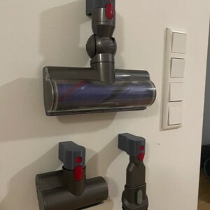 Puede incluir: Tres accesorios de aspiradora grises y rojos montados en una pared blanca. Los accesorios son un cabezal de cepillo motorizado, una boquilla para rendijas y un cepillo para el polvo.