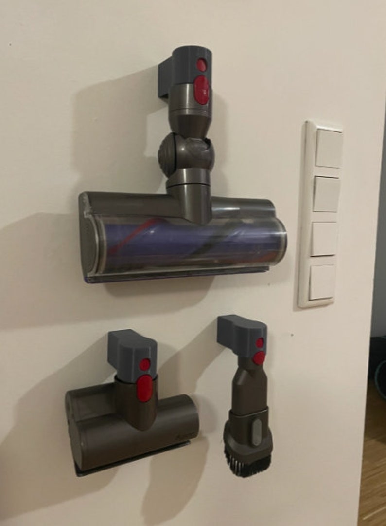Puede incluir: Tres accesorios de aspiradora grises y rojos montados en una pared blanca. Los accesorios son un cabezal de cepillo motorizado, una boquilla para rendijas y un cepillo para el polvo.