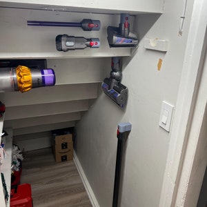 Puede incluir: Una pared blanca con una variedad de accesorios de aspiradora Dyson montados en ella. Los accesorios incluyen una boquilla para rendijas, un cepillo para polvo, un mini cabezal motorizado y una varilla de extensi&oacute;n larga.