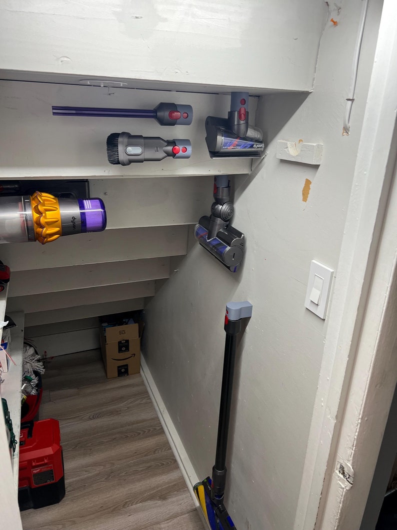 Puede incluir: Una pared blanca con una variedad de accesorios de aspiradora Dyson montados en ella. Los accesorios incluyen una boquilla para rendijas, un cepillo para polvo, un mini cabezal motorizado y una varilla de extensi&oacute;n larga.
