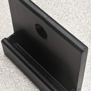 May include: A black plastic phone holder with a slot for a phone and a hole for a charging cable. The holder is designed to be mounted on a wall.