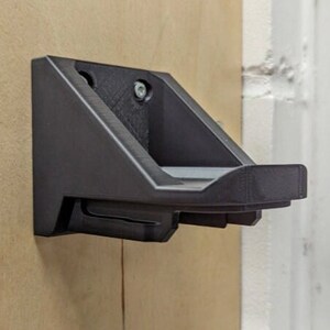 Pode incluir: Um suporte de parede de plástico preto com uma prateleira curva para segurar objetos. O suporte está fixado a uma parede de cor marrom claro.