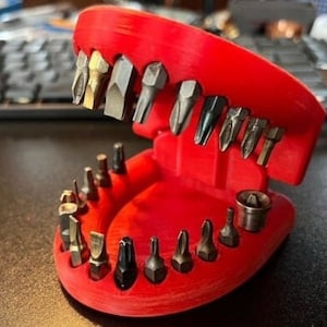 May include: A red plastic tool holder shaped like a mouth with a variety of different sized screwdriver bits. The holder has a row of 15 bits on the bottom and 8 bits on the top.