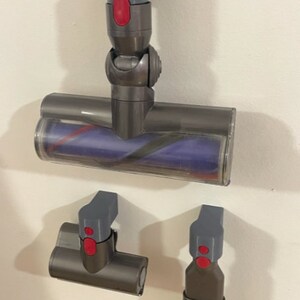 Puede incluir: Tres accesorios de aspiradora grises y rojos montados en una pared blanca. El accesorio superior es un cabezal de cepillo motorizado con una cubierta de pl&aacute;stico transparente. El accesorio del medio es una boquilla para ranuras. El accesorio inferior es un cepillo para polvo.