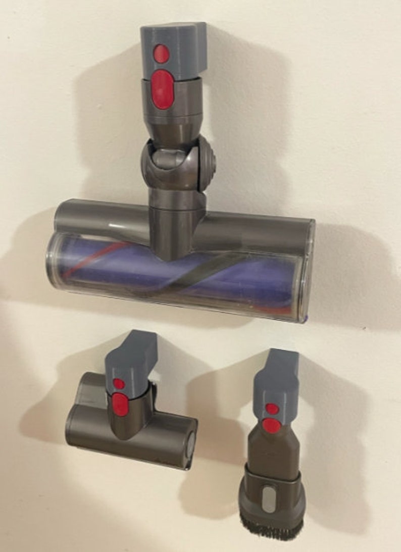 Puede incluir: Tres accesorios de aspiradora grises y rojos montados en una pared blanca. El accesorio superior es un cabezal de cepillo motorizado con una cubierta de pl&aacute;stico transparente. El accesorio del medio es una boquilla para ranuras. El accesorio inferior es un cepillo para polvo.