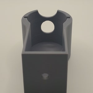 Puede incluir: Un soporte de pared de pl&aacute;stico gris impreso en 3D con una abertura circular para un dispositivo.