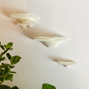 May include: Three white ceramic wall shelves shaped like mushrooms, mounted on a white wall. The shelves are arranged in a staggered pattern, with the largest shelf at the top and the smallest shelf at the bottom.