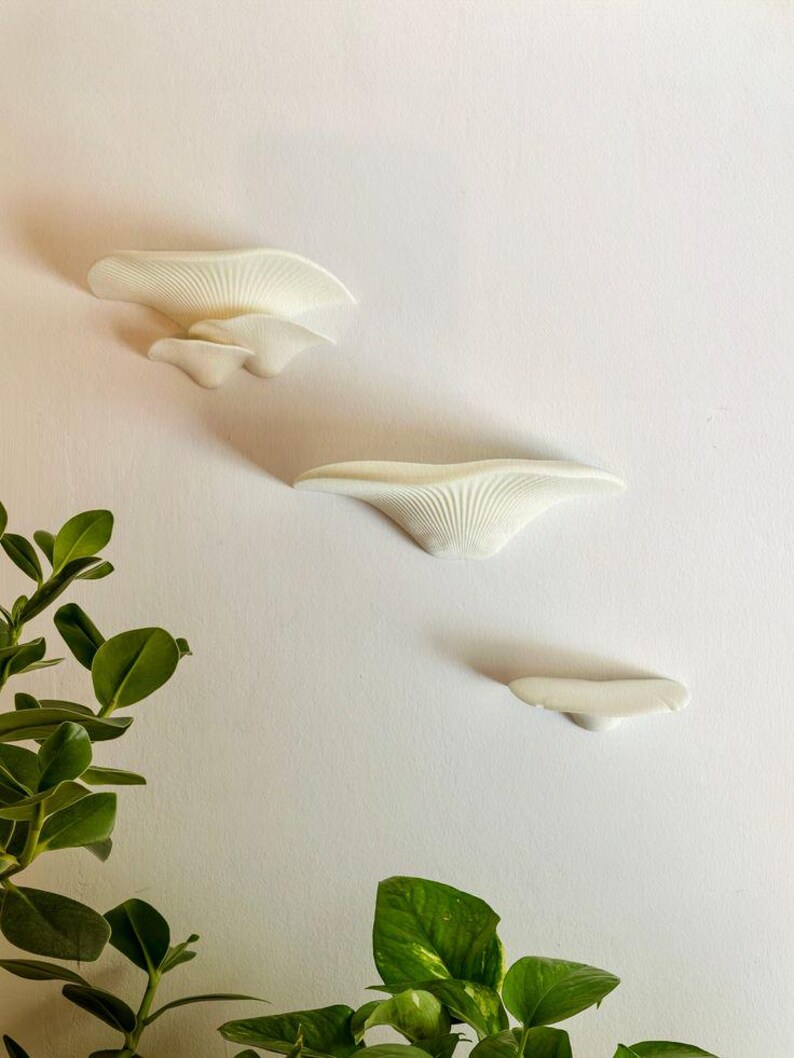 May include: Three white ceramic wall shelves shaped like mushrooms, mounted on a white wall. The shelves are arranged in a staggered pattern, with the largest shelf at the top and the smallest shelf at the bottom.