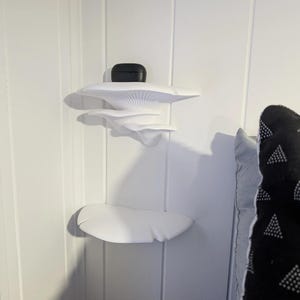 May include: White wall-mounted shelves shaped like mushrooms. The shelves are in a corner of a room and have a black wireless earbud case on the top shelf.