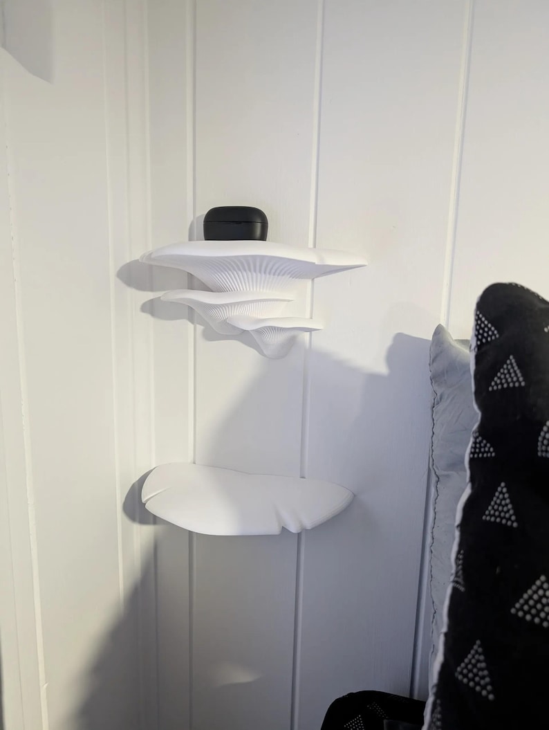 May include: White wall-mounted shelves shaped like mushrooms. The shelves are in a corner of a room and have a black wireless earbud case on the top shelf.