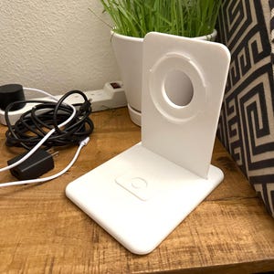 Op de afbeelding: Een witte draadloze oplaadstandaard met een cirkelvormig oplaadgedeelte en een rechthoekige basis. Een zwarte voedingsadapter en een zwarte oplaadkabel zijn zichtbaar op een houten oppervlak. Een kleine pot met gras staat op de achtergrond.