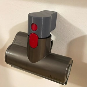 May include: A gray and brown Dyson vacuum cleaner attachment with a red button and the word "Dyson" printed on the side. The attachment is mounted on a wall.