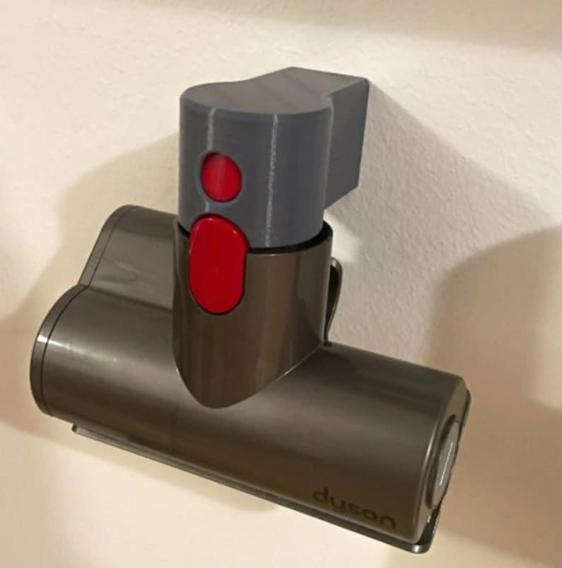 Puede incluir: Un accesorio de aspiradora Dyson de color gris y marr&oacute;n con un bot&oacute;n rojo y la palabra "Dyson" impresa en el lateral. El accesorio est&aacute; montado en una pared.