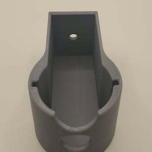 Puede incluir: Un soporte de pared de pl&aacute;stico gris con una abertura circular y un peque&ntilde;o agujero en la parte superior. El soporte est&aacute; dise&ntilde;ado para sujetar un objeto peque&ntilde;o, como un tel&eacute;fono o un altavoz.