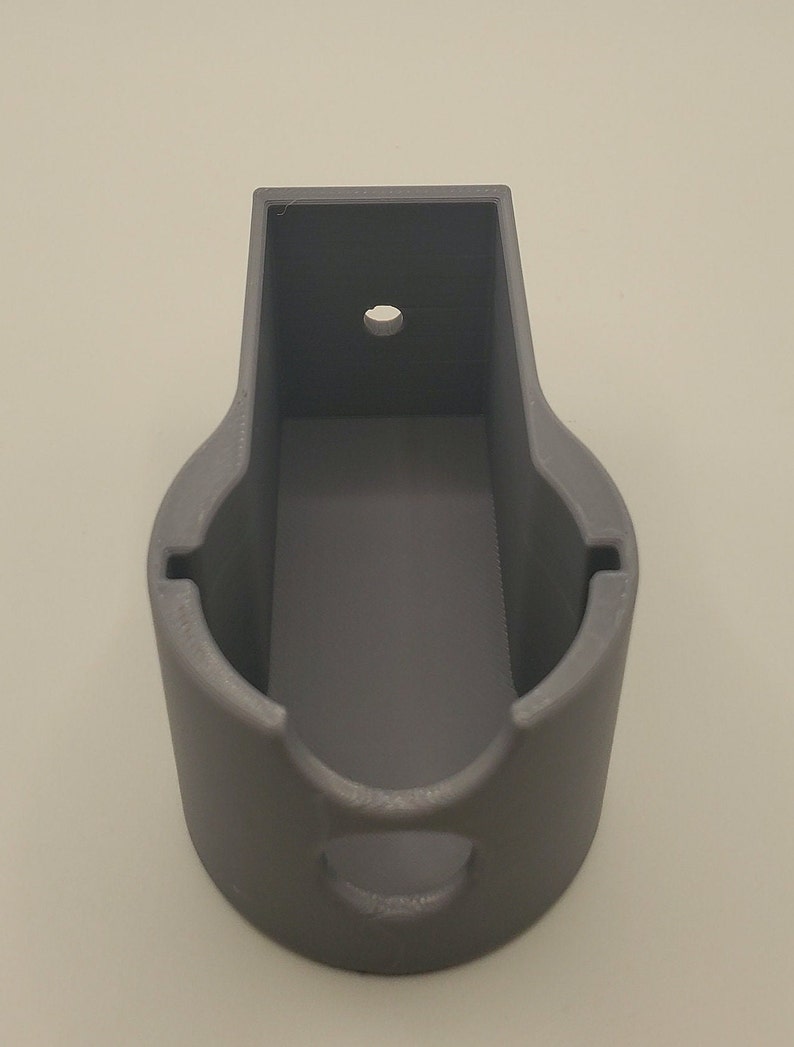 Puede incluir: Un soporte de pared de pl&aacute;stico gris con una abertura circular y un peque&ntilde;o agujero en la parte superior. El soporte est&aacute; dise&ntilde;ado para sujetar un objeto peque&ntilde;o, como un tel&eacute;fono o un altavoz.