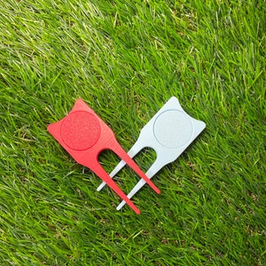 May include: Two golf ball retrievers, one red and one light blue, with a rounded top and three prongs. The retrievers are lying on a green grassy surface.