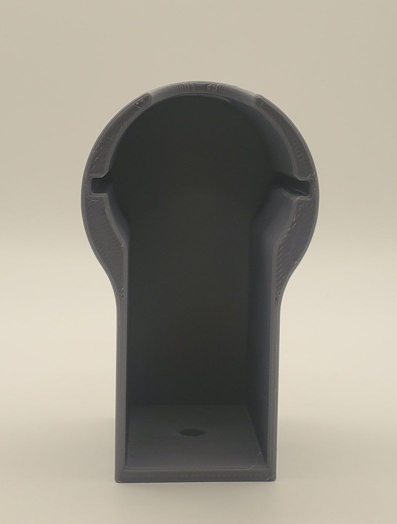 Puede incluir: Un soporte de pl&aacute;stico gris impreso en 3D con una abertura redonda en la parte superior y una base rectangular. El soporte tiene un peque&ntilde;o agujero en la parte inferior.