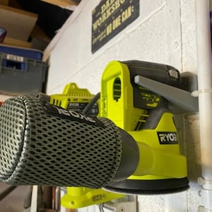 Può includere: Due utensili elettrici Ryobi montati su una parete bianca con un cartello nero che recita "Officina di papà - Nessuno può". Gli utensili sono una levigatrice verde e nera e un trapano verde e nero. La levigatrice ha una copertura in rete nera sul davanti.