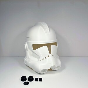 Könnte beinhalten: Ein weißer Clone Trooper Helm aus Kunststoff mit T-förmigem Visier und Griff oben. Der Helm hat detaillierte Gesichtsmerkmale und wird von mehreren kleinen, schwarzen Komponenten begleitet.