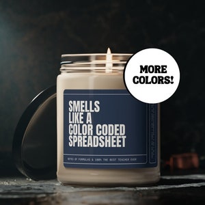 May include: A beige candle with a blue label that reads "Smells like a color coded spreadsheet" and "Notes of formulas & 100% the best teacher ever". A white sticker on the side reads "More Colors!"