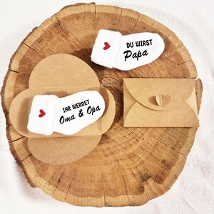 May include: Two white baby socks with red hearts. One sock says "Du wirst Papa" and the other says "Ihr werdet Oma & Opa". The socks are in brown paper envelopes.