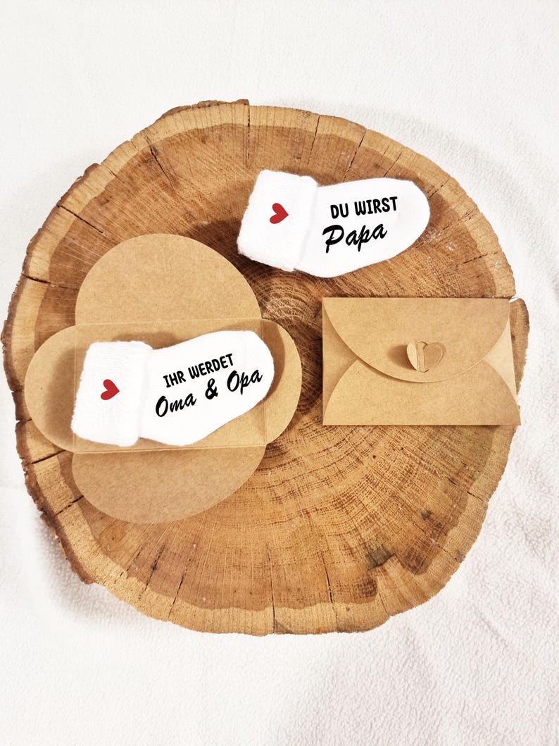May include: Two white baby socks with red hearts. One sock says "Du wirst Papa" and the other says "Ihr werdet Oma & Opa". The socks are in brown paper envelopes.
