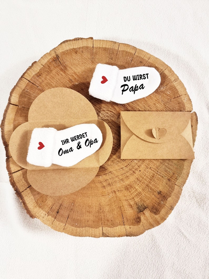 May include: Two white baby socks with red hearts and black text. One sock says "Du wirst Papa" and the other says "Ihr werdet Oma & Opa". The socks are in a clear plastic box with a brown cardboard backing. The box is on a brown wooden surface.