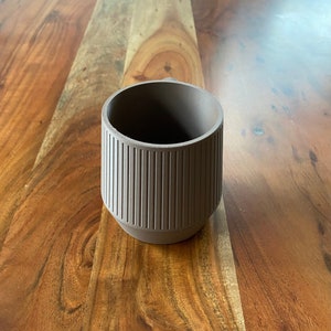 Könnte beinhalten: Eine braune Keramiktasse mit geripptem Design. Die Tasse steht auf einer Holzoberfläche.