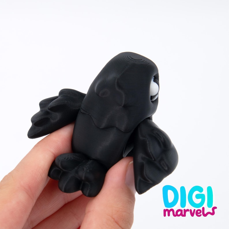 Peut inclure: Une petite figurine d'oiseau imprim&eacute;e en 3D, de couleur noir mat, avec des ailes stylis&eacute;es et un seul &oelig;il blanc visible. La surface pr&eacute;sente de fines lignes horizontales dues &agrave; l'impression. Le texte DIGI marvels est &eacute;crit en bleu et rose.