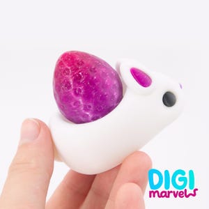 May include: A white bunny-shaped toy holding a purple and pink egg-shaped object. The bunny has black eyes and pink inner ears. The toy is held in a hand against a white background. The text "DIGI marvels" is in the bottom right corner.