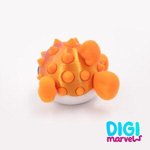 May include: A 3D printed pufferfish figurine with a metallic orange upper body covered in rounded spikes and a smooth white underbelly. The figure features orange fins and the text DIGI marvels printed in blue and magenta in the bottom corner.