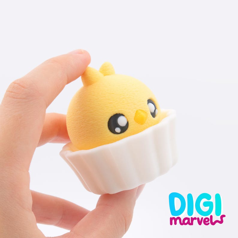 May include: A yellow chick-shaped toy with large eyes and a small beak, set in a white cupcake liner. The toy is held in a hand. The Digi Marvels logo is visible in the bottom right corner.