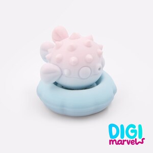 May include: A light pink pufferfish figurine with small spikes on its body, resting inside a light blue circular floatation ring. The object features a smooth matte finish and rounded fins. The text DIGI marvels is printed in the bottom right corner.