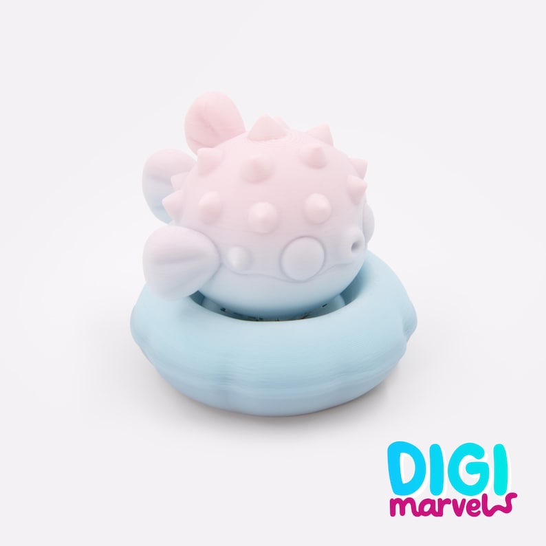 May include: A light pink pufferfish figurine with small spikes on its body, resting inside a light blue circular floatation ring. The object features a smooth matte finish and rounded fins. The text DIGI marvels is printed in the bottom right corner.