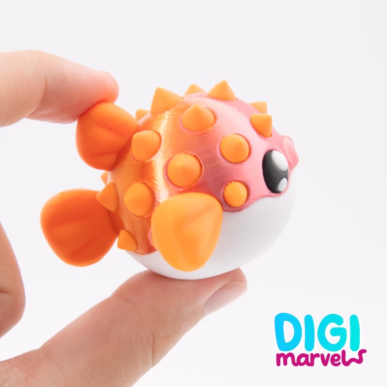 May include: A small, spherical pufferfish figurine featuring a gradient orange and pink body with protruding orange spikes, orange fins, and a white underbelly. The fish has a large black eye with white highlights and the text DIGI marvels.