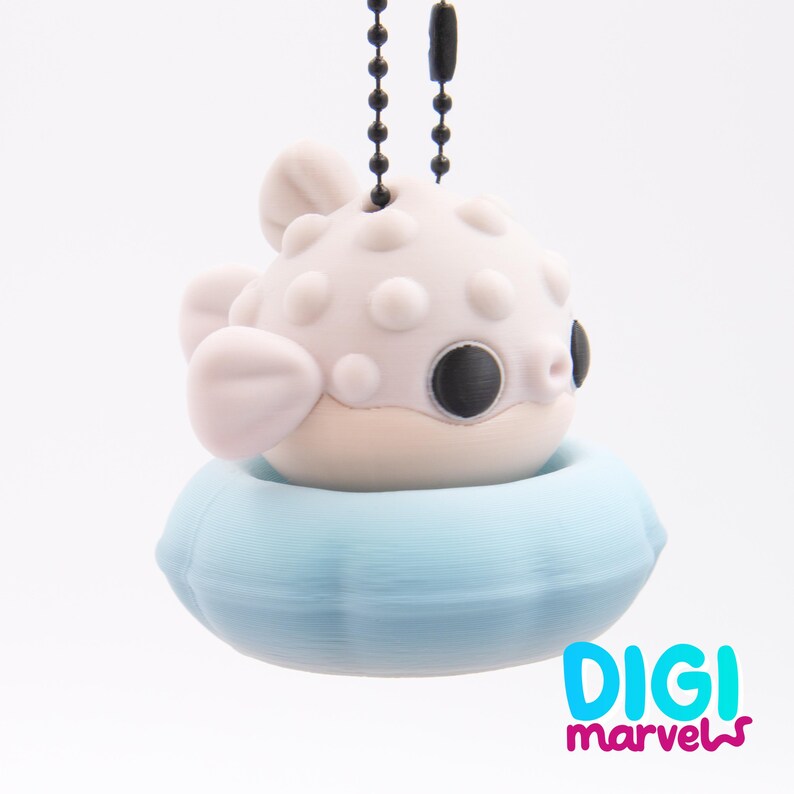 May include: A light pink pufferfish figurine with large black eyes, attached to a black ball chain, resting inside a light blue circular floatation ring. The text DIGI marvels is printed in blue and magenta in the bottom right corner.