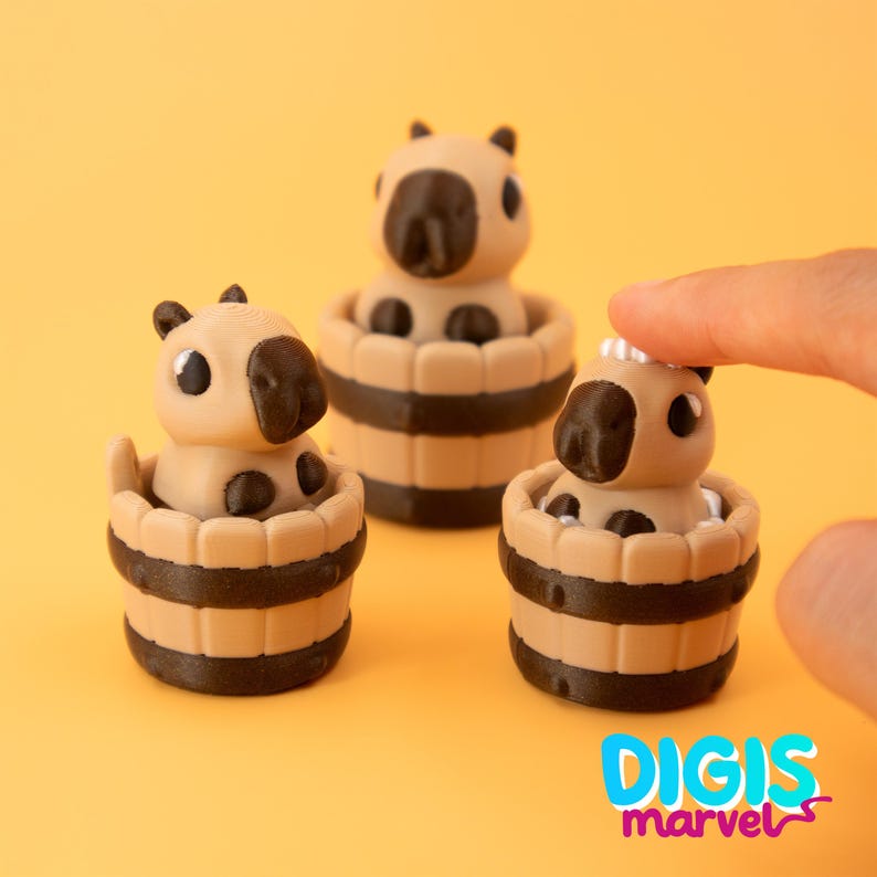 May include: Three miniature capybara figurines in wooden tubs. The capybaras are tan with black details, and the tubs are tan with dark brown stripes. The background is a solid yellow-orange color. The logo "DIGIS marvel" is in the bottom right corner.