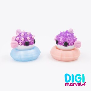 May include: Two small pufferfish figurines with purple bodies and pink spikes, each resting in a circular float. One float is light blue and the other is peach-colored. The text DIGI marvels is printed in the bottom right corner in blue and pink.