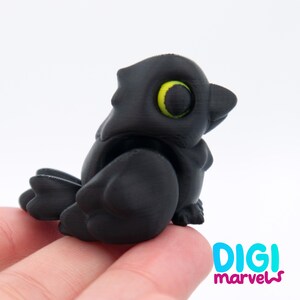 Peut inclure: Une petite figurine d'oiseau noir mat avec un corps arrondi et stylis&eacute;, dot&eacute;e de grands yeux pro&eacute;minents aux iris jaune vif. L'oiseau pr&eacute;sente une surface lisse et unie, repr&eacute;sent&eacute; en position assise avec les ailes repli&eacute;es.