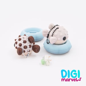 May include: Two pufferfish-shaped keycaps with ball chains, one light gray and one white with brown spikes, each resting in a light blue circular base. A single mechanical keyboard switch with a light green stem sits in the center. Text reads DIGI marvels.