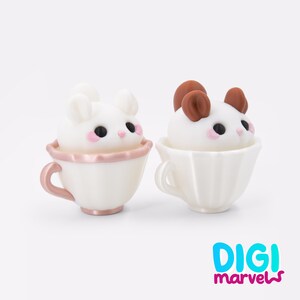 Puede incluir: Dos peque&ntilde;as figuras de criaturas blancas con ojos negros y mejillas rosadas, cada una sentada dentro de una taza de t&eacute; estriada. La taza izquierda es blanca con asa y borde rosa met&aacute;lico, la derecha es blanca. Se ve el texto DIGI marvels.