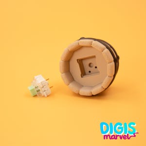 May include: A beige and brown keyboard keycap with a square cutout, next to a white and green mechanical keyboard switch. The keycap has a circular design with a dark brown border. The background is a solid orange color, with the text "DIGIS marvel" in the bottom right corner.