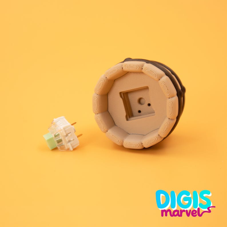 May include: A beige and brown keyboard keycap with a square cutout, next to a white and green mechanical keyboard switch. The keycap has a circular design with a dark brown border. The background is a solid orange color, with the text "DIGIS marvel" in the bottom right corner.