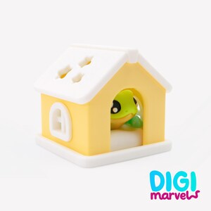 Pode incluir: Uma pequena estatueta em forma de casa amarela e branca com um telhado branco com recortes em forma de estrela e uma moldura de janela branca. Um sapo verde com grandes olhos pretos &eacute; vis&iacute;vel dentro. Texto DIGI marvels no canto inferior.