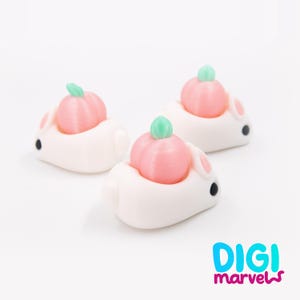 May include: Three white bunny figurines, each with a pink pumpkin on its back. The pumpkins have green stems. The bunnies have black eyes and pink inner ears. The figurines are on a white background.
