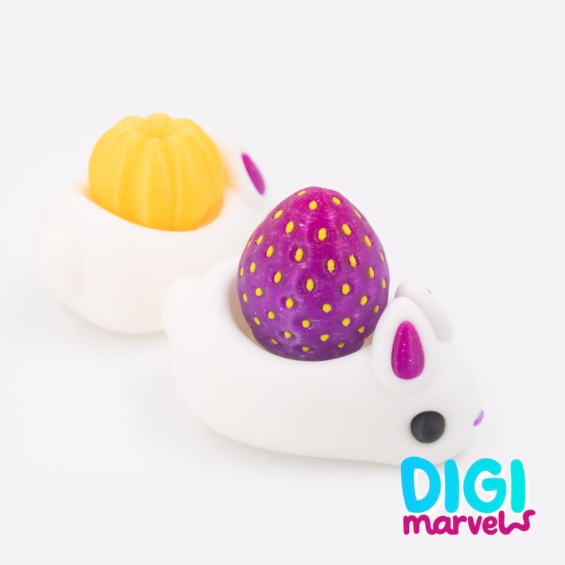 May include: Two white bunny-shaped holders, each cradling a colorful fruit-like object. One holds a yellow pumpkin, the other a purple strawberry with yellow dots. The bunnies have pink inner ears and black eyes. The image includes the text "DIGI marvels".