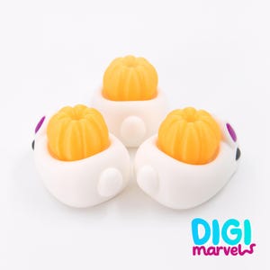 May include: Three white bunny-shaped figurines, each holding a small, bright orange pumpkin. The bunnies have purple accents on their ears and black eyes. The image includes the text "DIGI marvels" in blue and pink.