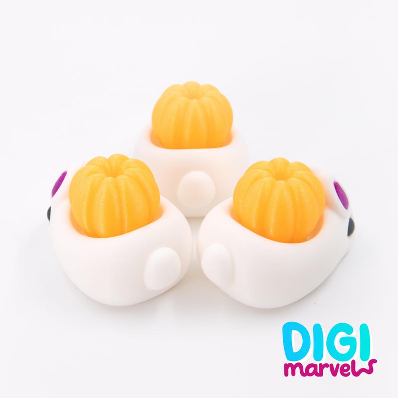 May include: Three white bunny-shaped figurines, each holding a small, bright orange pumpkin. The bunnies have purple accents on their ears and black eyes. The image includes the text "DIGI marvels" in blue and pink.
