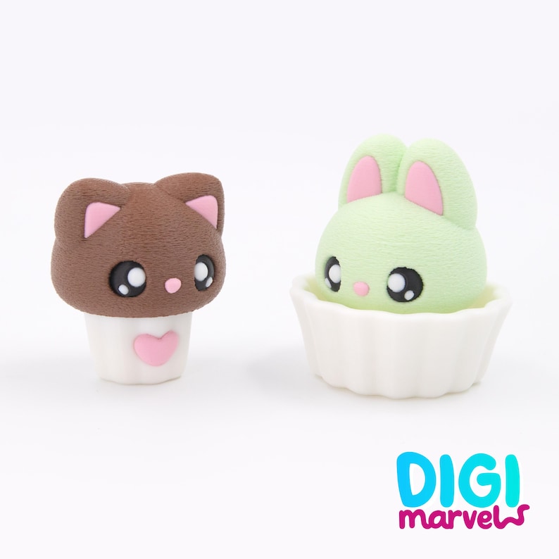 May include: Two cute figurines: a brown cat with pink details and a green bunny with pink ears. The cat has a white base with a pink heart, and the bunny sits in a white cupcake case. The Digi Marvels logo is in the bottom right.